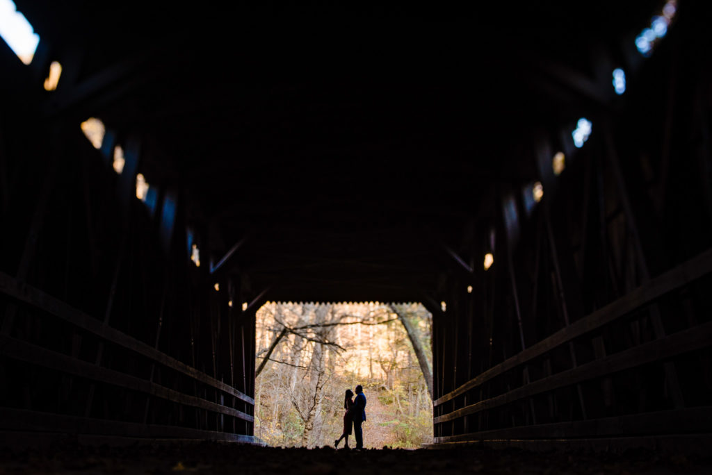 Silholutte Photo of couple in Thomas Mill Covered Bridge during their engagement session at the Forbidden Drive pathway. Kristin goes in for a kiss.  